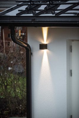 - LedStore Moderni LED-ulkovalaisin valkoisella seinällä, valaistus ylös/alas; siinä on musta kattokouru, metallinen syöksyputki, lehdettömät pensaat taustalla.