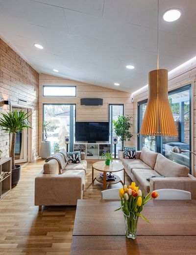 Soggiorno moderno con grandi finestre, divani beige, TV, piante, tavolo in legno con vasi di tulipani, pareti in pannelli di legno - illuminato con un'elegante illuminazione a LED.