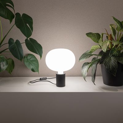 Moderna lampada da tavolo a LED con paralume rotondo bianco su mensola, circondata da piante verdi in vaso per un'elegante illuminazione domestica.