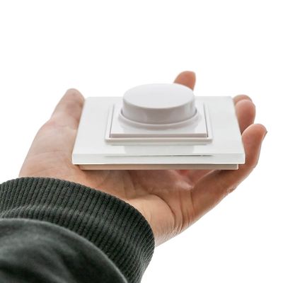 Una persona tiene in mano un interruttore dimmer bianco su una manica verde scuro, esaltando il controllo dell'illuminazione a LED su uno sfondo bianco.