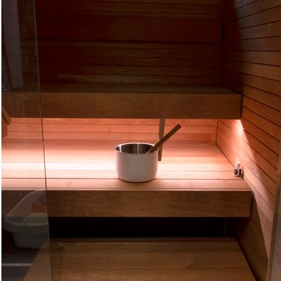 Interno di sauna in legno con secchio e paletta in metallo sulle panche; l'illuminazione calda a LED presenta le ragioni del legno sulle superfici di seduta.