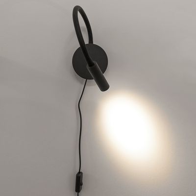 Moderna lampada da parete nera con collo regolabile e base rotonda; emette una luce bianca morbida su una parete liscia. Ideale per un'illuminazione domestica elegante e funzionale.