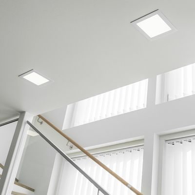 Luminose luci quadrate da incasso a soffitto illuminano una scala moderna con balaustra in vetro e grandi finestre con tende verticali.
