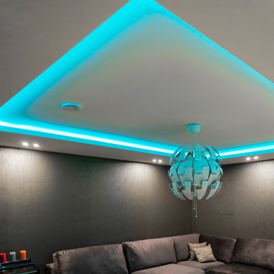 Un soggiorno moderno con un divano grigio, luci LED blu-verdi a soffitto, una luce a sospensione circolare e un tavolino con candele vivaci per l'illuminazione d'ambiente.