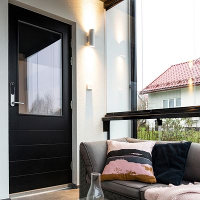 Moderna veranda con illuminazione a LED ad alta efficienza energetica, porta nera, divano grigio con cuscini colorati, pareti di vetro che mostrano la casa con un tetto rosso vivace.