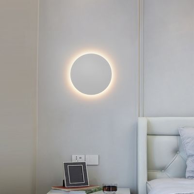 Una moderna applique circolare illumina una camera da letto minimalista con pareti grigio chiaro, una testiera del letto bianca e un'elegante decorazione sul comodino. Negozio di illuminazione a LED.