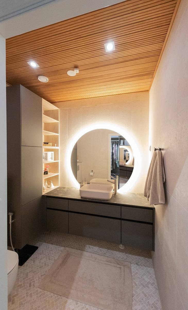 Un bagno moderno con illuminazione a LED di LedStore.fi: uno specchio circolare retroilluminato a LED sopra il lavandino crea un'atmosfera elegante. Il bagno è caratterizzato da armadietti scuri, soffitti in legno caldo e pareti chiare. Gli asciugamani sono posizionati sul lato destro e le mensole ti aspettano sul lato sinistro. Il pavimento è ricoperto da un tappeto beige che contribuisce all'effetto complessivo.   