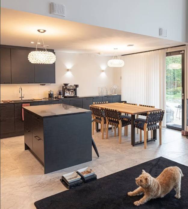 Una cucina e una sala da pranzo moderne con armadietti neri, un tavolo da pranzo in legno e una comoda illuminazione LED di LedStore.fi creano un'atmosfera accogliente. Un gatto che si stiracchia su un tappeto nero osserva l'insieme accogliente. 
