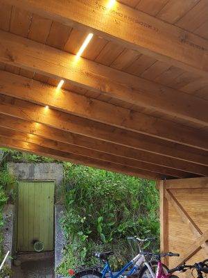 Tettoia in legno, illuminata da una striscia LED che corre tra il tetto e i parapetti del tetto. Biciclette sotto la tettoia e una porta verde sullo sfondo.