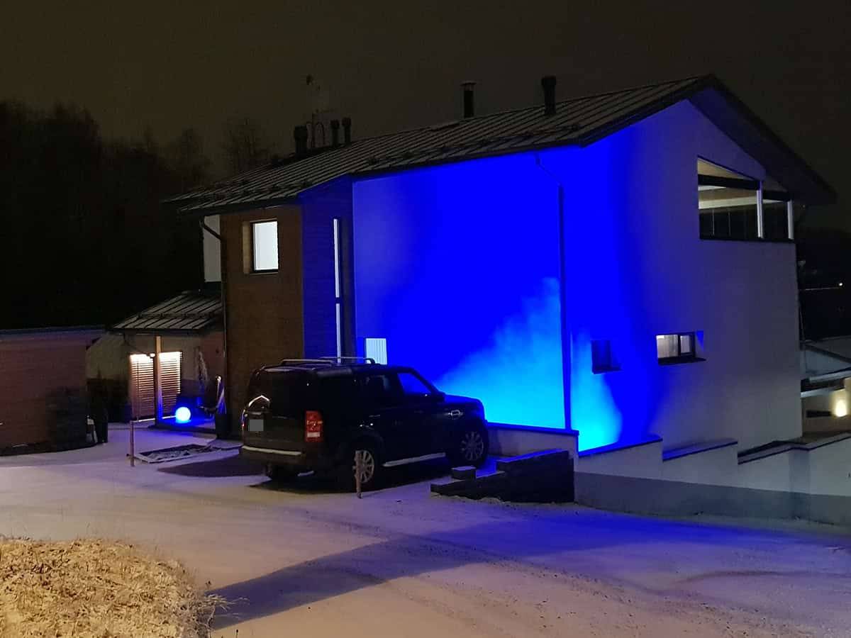 La facciata di una casa moderna illuminata di notte da luci LED blu. Un'auto nel cortile e un leggero manto di neve sul terreno.