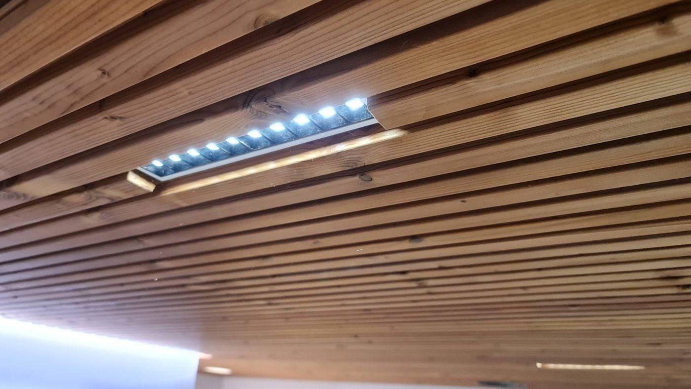 Illuminazione a soffitto a strisce e soffitto a strisce a basso costo Il soffitto in legno a doghe parallele è dotato di illuminazione LED a incasso. I LED sono parzialmente visibili attraverso un'apertura rettangolare, creando un sottile bagliore sulla superficie naturale del legno.