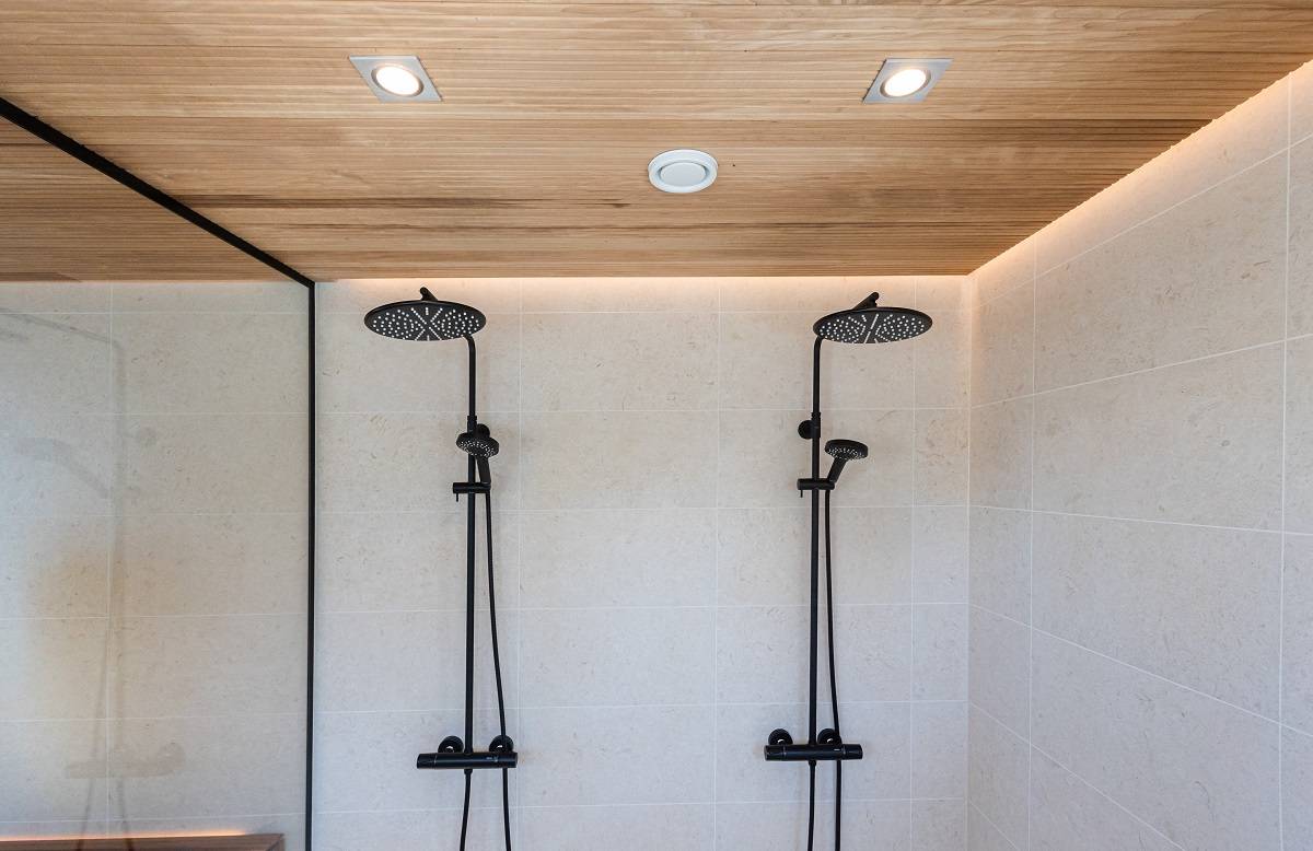 Il moderno bagno con doccia è dotato di due soffioni neri e di piastrelle color crema. Il soffitto rivestito in legno, accentuato da luci LED a incasso di LedStore.fi, crea un look minimalista ed elegante. La zona doccia è separata dal resto dello spazio da una parete esterna in vetro.  