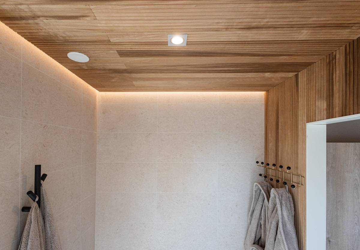 LedStore.fi offre una soluzione per il bagno moderno con un'illuminazione LED moderna. Le pareti piastrellate beige, il soffitto in legno e l'illuminazione LED a incasso creano un look fresco e moderno. Morbidi asciugamani grigi appesi a ganci neri armonizzano l'interno minimalista.  