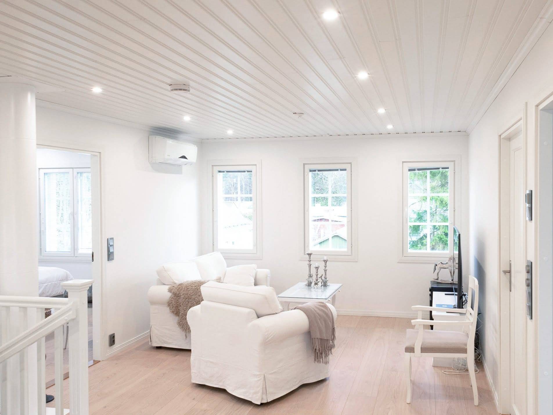Soggiorno moderno con illuminazione a LED, poltrone bianche, tavolino e TV; la luce naturale proviene dalle ampie finestre e accentua il pavimento in legno.