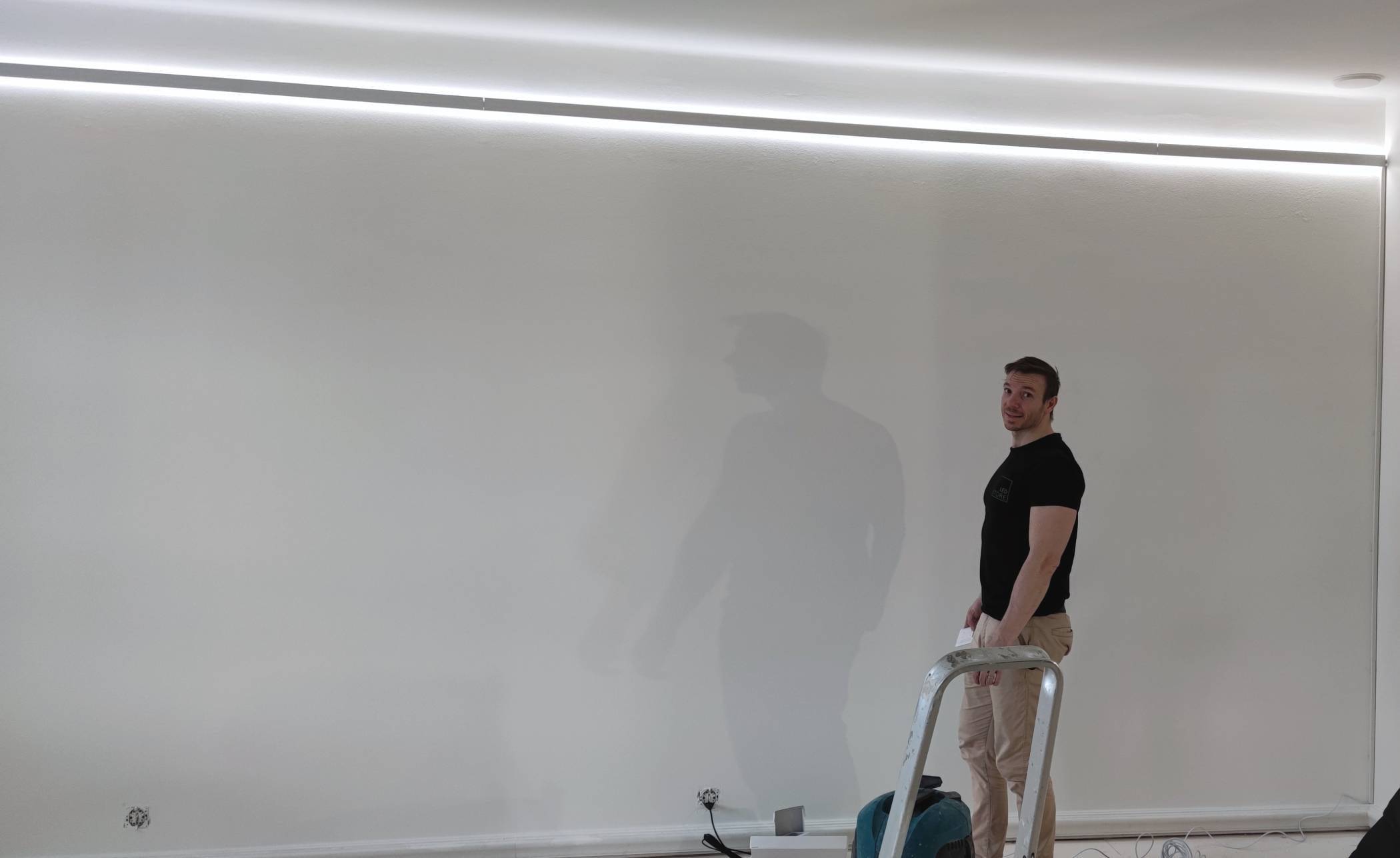 Una persona su una scala con un attrezzo, con un'illuminazione LED soffusa dall'alto, su una parete bianca. Cavi e attrezzature sparsi sul pavimento con un'illuminazione LED da negozio.