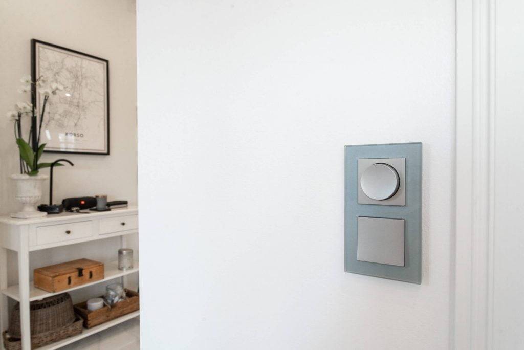 Interruttore LED moderno con due pulsanti su una parete bianca; console bianca con orchidea in vaso e mappa incorniciata in una stanza minimalista.