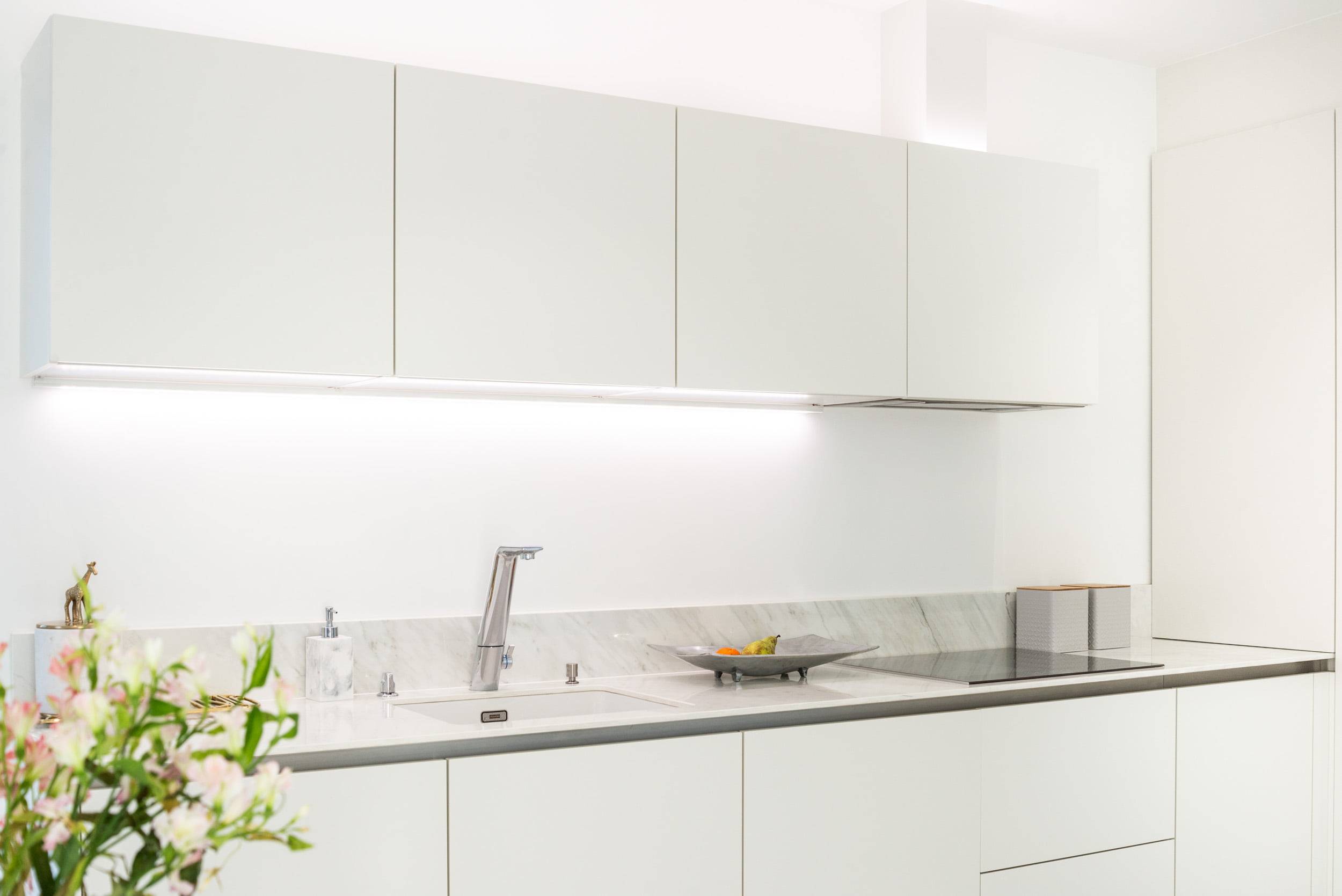 Una cucina elegante con armadietti bianchi e puliti, un moderno piano di lavoro in marmo e un rubinetto in acciaio inox. L'illuminazione LED sotto il banco accentua l'aspetto dello spazio di lavoro. Una pianta decorativa e una fruttiera aggiungono ulteriore fascino al piano di lavoro. Puoi trovare le giuste soluzioni di illuminazione LED su LedStore.fi, dove potrai trovare prodotti LED di qualità per completare il look della tua cucina.
