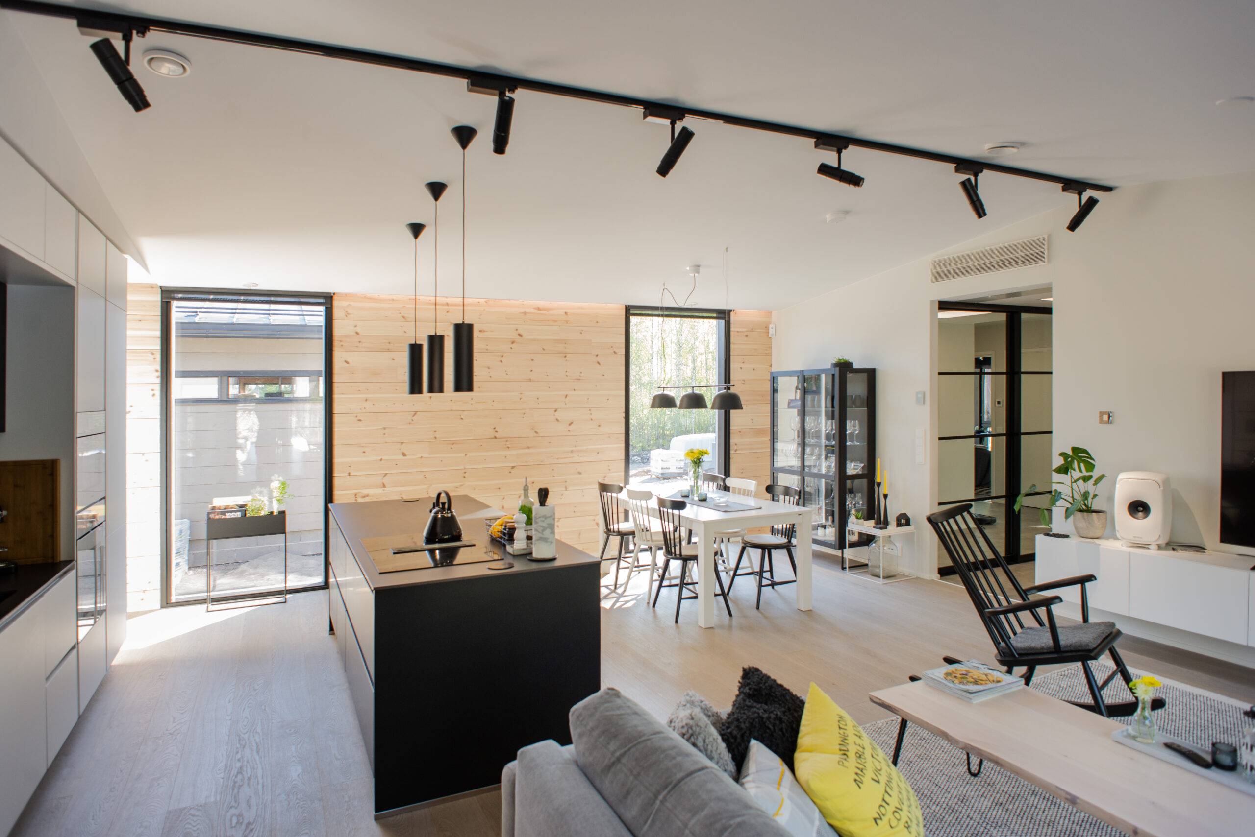 Cucina e soggiorno moderni a pianta aperta con dettagli in legno e mobili neri per un look elegante. L'illuminazione interna di alta qualità di LedStore.fi crea una grande atmosfera e le ampie finestre lasciano entrare la luce naturale.