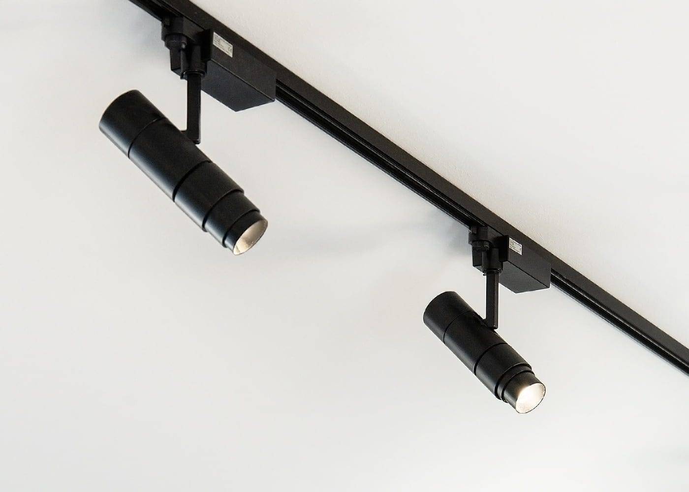 La configurazione minimalista con due binari LED cilindrici neri su un tetto bianco, montati su un binario nero, mette in mostra un design di illuminazione moderno e angoli regolabili.