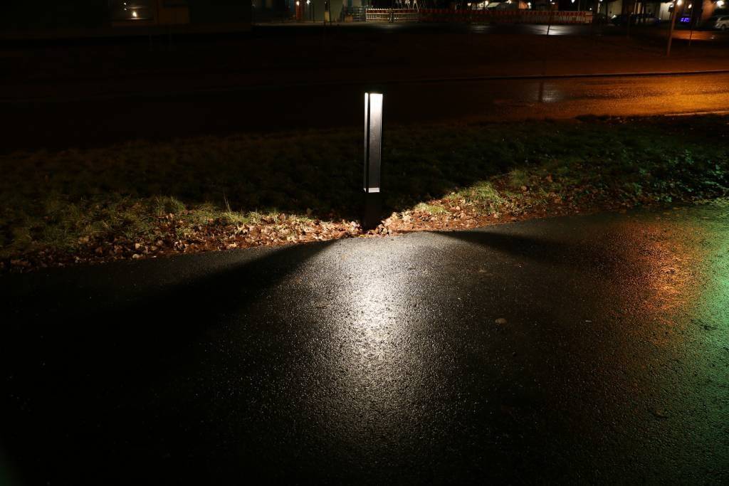 Un lampione a LED che illumina una strada bagnata con motivi triangolari delimitati dall'erba; uno sfondo urbano con luci lontane.