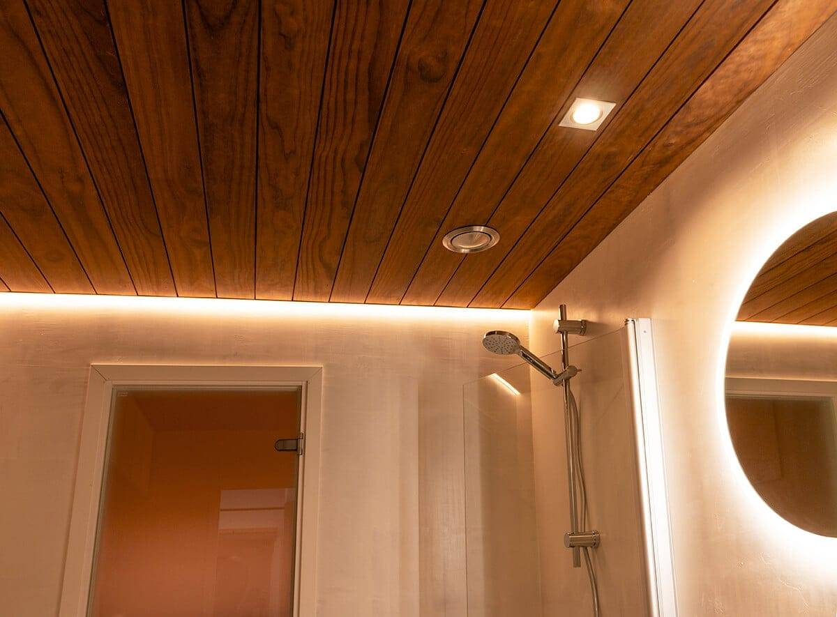 Bagno con soffitto in legno e illuminazione ambientale a LED, soffione moderno, luce a spot e specchio circolare a LED; porta a sinistra.
