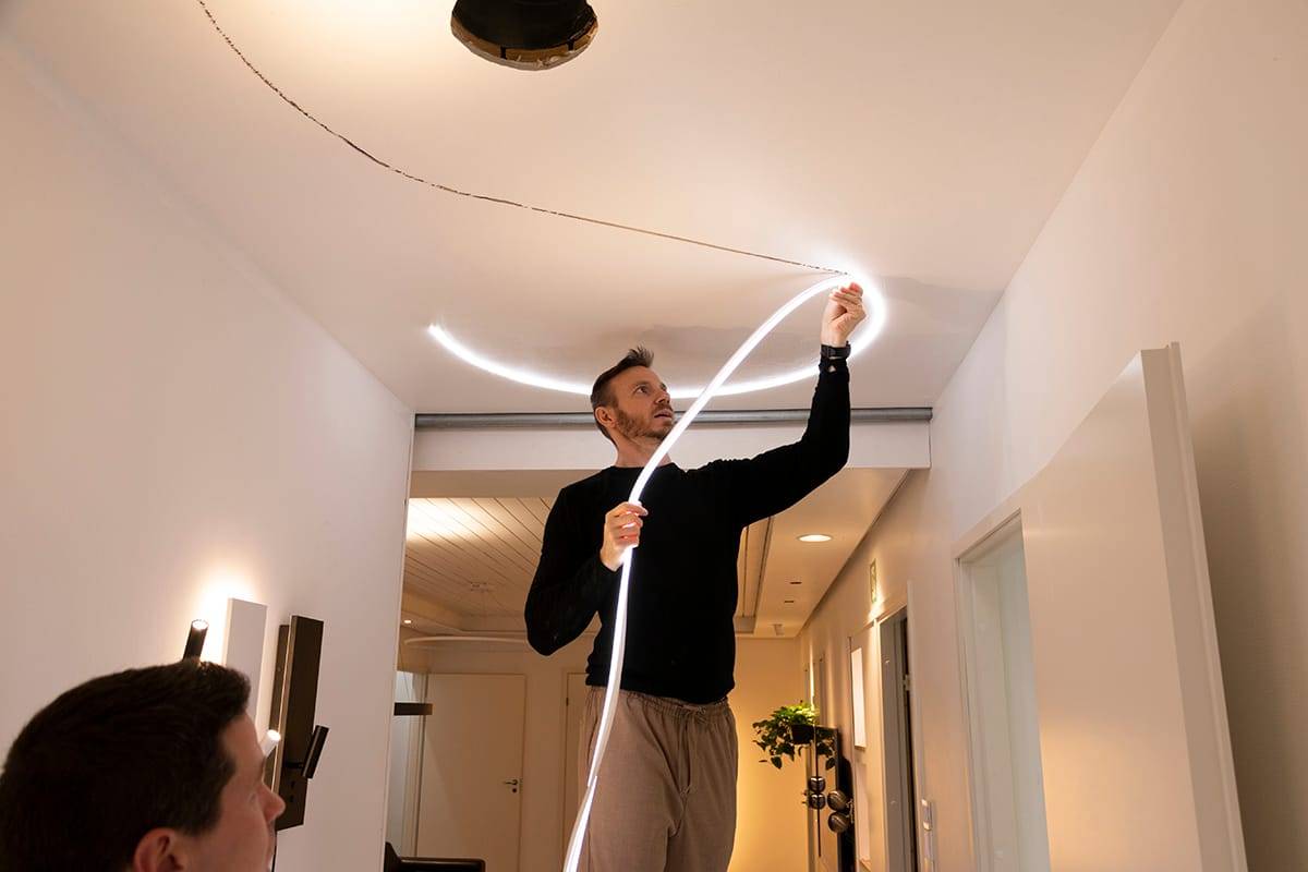 Una persona attacca una striscia di luce LED flessibile a un soffitto moderno, migliorando l'illuminazione della stanza. Maglione nero, pantaloni beige; un'altra persona in primo piano, sfocata.