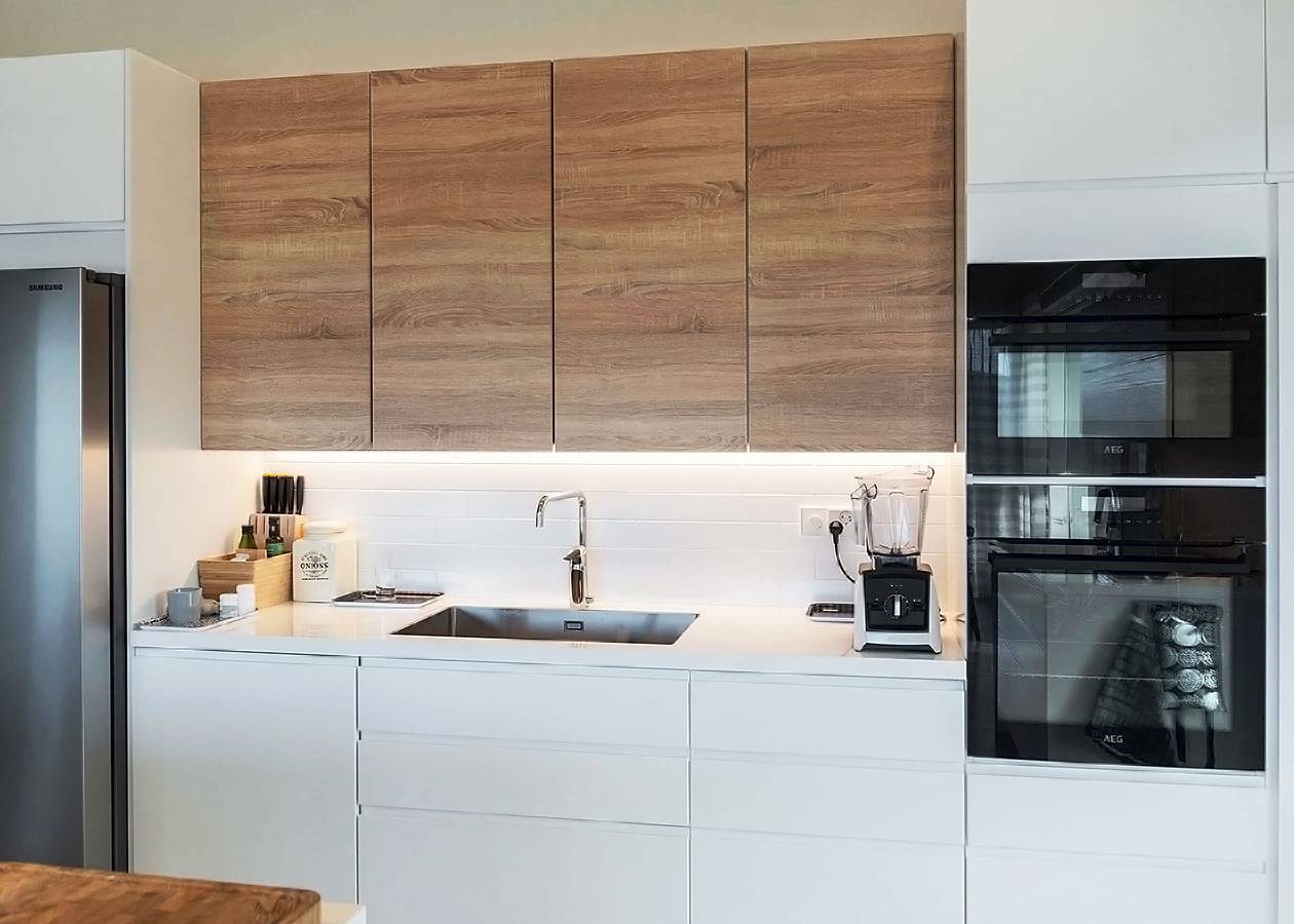 Cucina moderna con armadietti superiori in legno, piani di lavoro bianchi illuminati a LED, lavello in acciaio inox con rubinetto cromato, doppio forno ed elettrodomestici da appoggio.