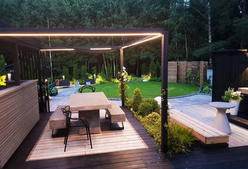 Moderna terrazza esterna con pergola con illuminazione a LED incorporata, tavolo in cemento, panche con terrazza in legno; giardino rigoglioso e prato curato.