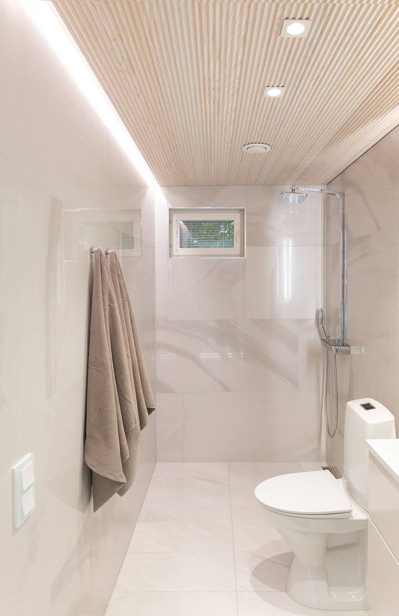 Un bagno moderno e minimalista con pareti beige chiaro e soffitto piastrellato in legno. Un portasciugamani a parete con due asciugamani, una piccola finestra e un'area doccia separata da un divisorio in vetro aggiungono eleganza allo spazio. Le plafoniere a LED integrate garantiscono un'illuminazione efficace. LedStore.fi: Scopri le soluzioni di illuminazione a LED perfette per interni eleganti e senza tempo.   