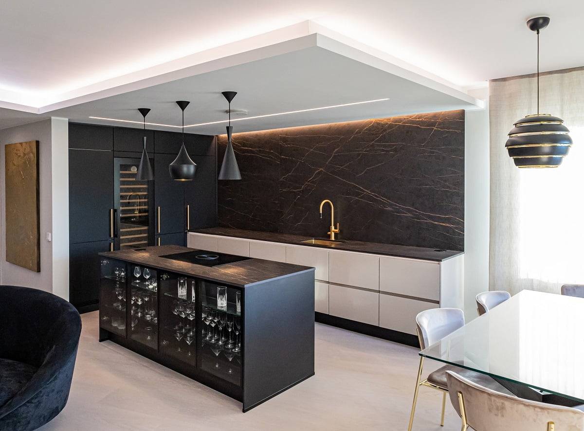 Una cucina moderna con piastrelle bianche e nere, piastrelle bianche, un piano di lavoro in marmo nero e un rubinetto color oro. Le luci a sospensione illuminano un'isola nera con vetreria che perde. Un tavolo da pranzo adiacente in vetro ha sedie imbottite e sopra di esso brilla una suggestiva luce a LED a sospensione nera e oro. Dai un'occhiata all'ampia gamma di luci LED di LedStore.fi per creare un look simile a casa tua!
