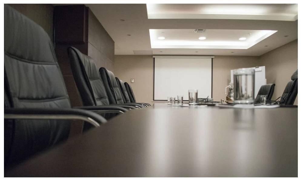 Sala conferenze con grande tavolo scuro e sedie. Sul soffitto sono presenti diversi pannelli e faretti.  
