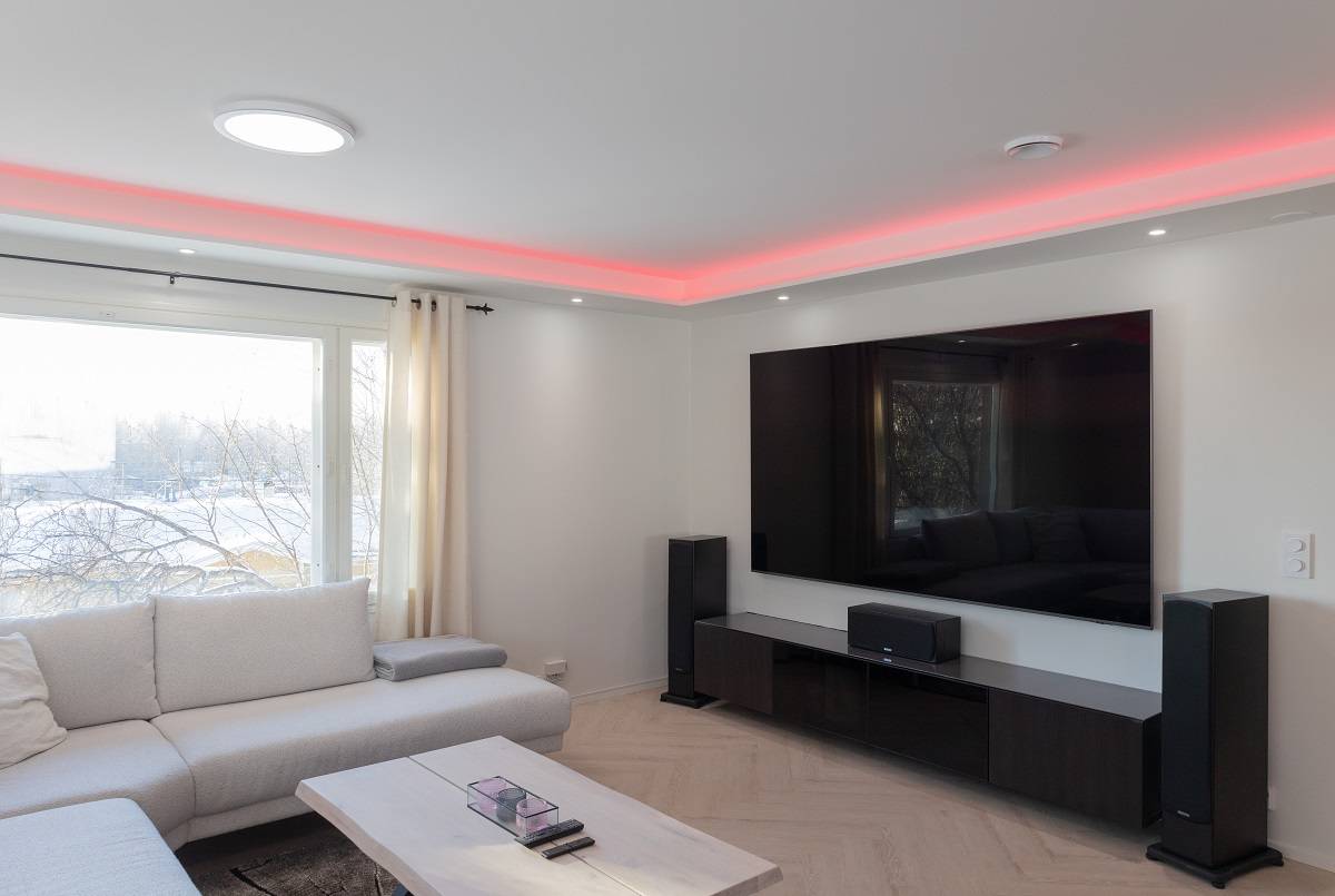 Un soggiorno moderno e ricco di atmosfere eleganti. Un ampio divano ti invita a rilassarti, mentre un tavolino in legno chiaro aggiunge calore allo spazio. Una TV a schermo piatto montata a parete con retroilluminazione a LED aggiunge un tocco moderno alla stanza. Amplifica la tua esperienza con altoparlanti da pavimento e goditi l'illuminazione d'accento a LED rossi sul soffitto. Un'ampia finestra fa entrare la luce naturale, rendendo l'ambiente spazioso e luminoso. Tutti questi elementi si combinano per creare l'ambiente perfetto con le versatili soluzioni di illuminazione LED di LedStore.fi.