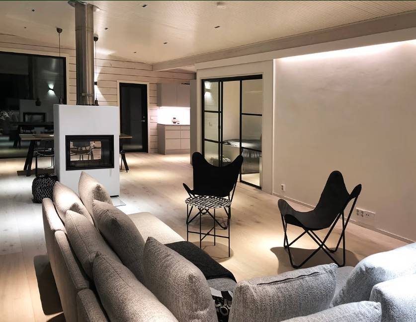 Un soggiorno moderno con un divano grigio, sedie nere, un caminetto e una cucina a vista sullo sfondo: l'atmosfera e lo stile sono creati dalla soluzione di illuminazione LED di alta qualità di LedStore.fi.