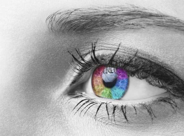 Primo piano di un occhio di colore grigio con iride iridescente simile a un LED in colori brillanti: rosso, blu, verde, viola; contrasto netto con pelle e ciglia monocromatiche.