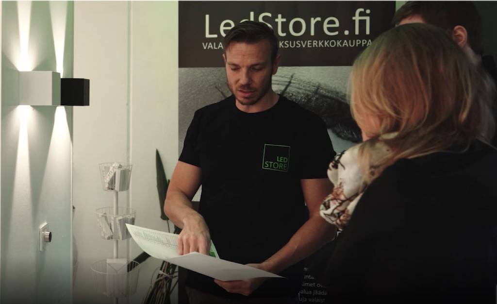 Un uomo con una maglietta nera di LED Store parla energicamente con una donna bionda, un'altra persona ascolta nelle vicinanze; sullo sfondo un'insegna "LedStore.fi" e luci LED luminose.
