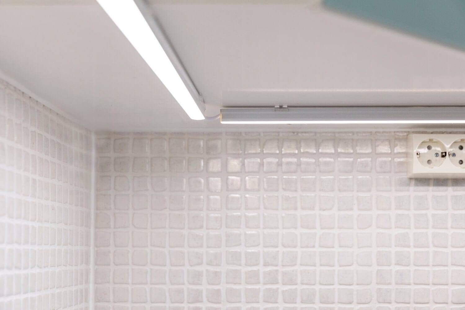 Un moderno paraschizzi da cucina con piastrelle bianche a texture, illuminato da una striscia LED lineare posizionata sotto il pensile; una luce soffusa mette in risalto la presa a muro.
