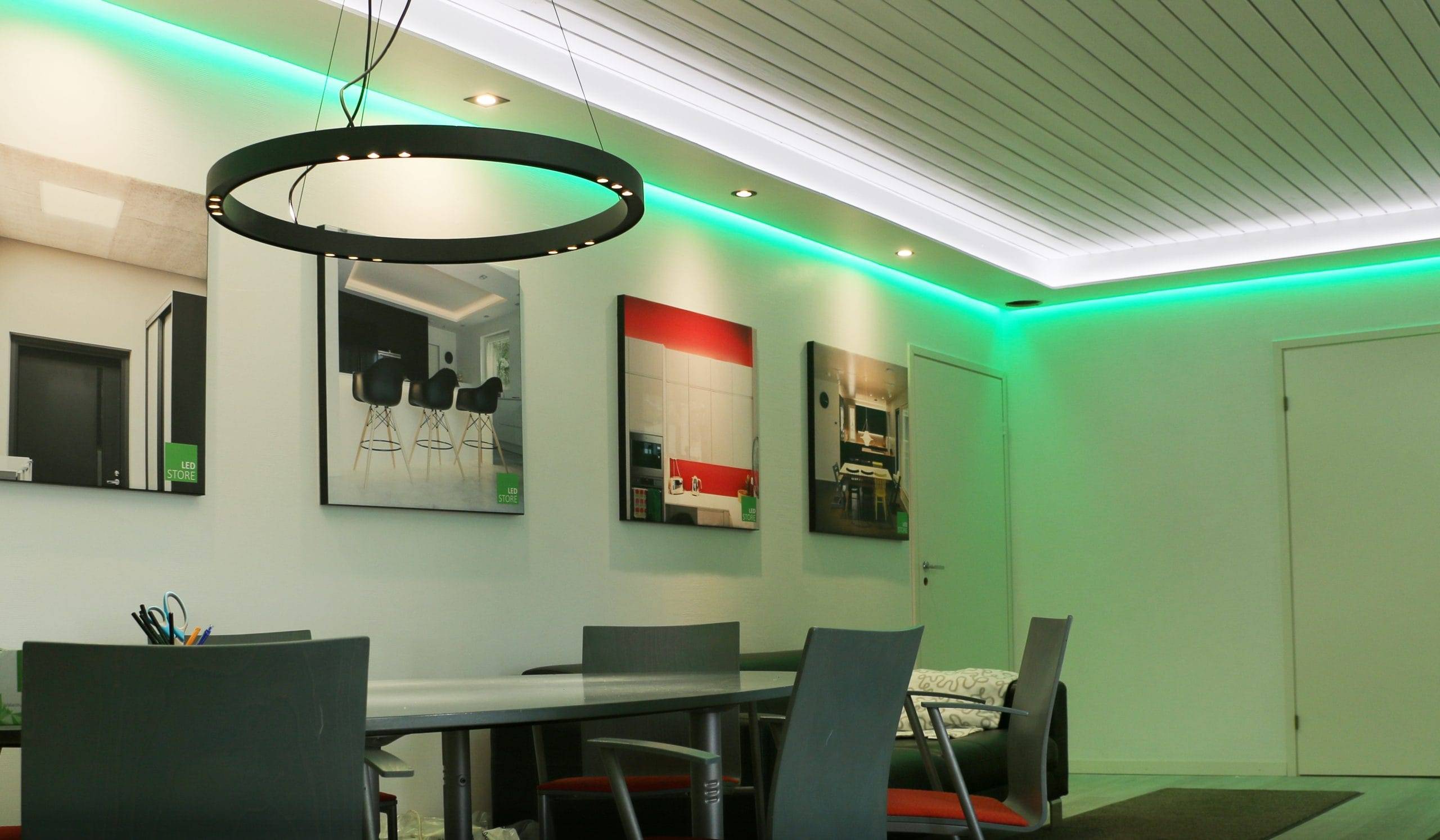 Una stanza moderna con una luce LED circolare nera, sedie grigie intorno a un lungo tavolo, opere d'arte incorniciate alla parete e illuminazione a soffitto con riflessi verdi per creare un'atmosfera elegante.