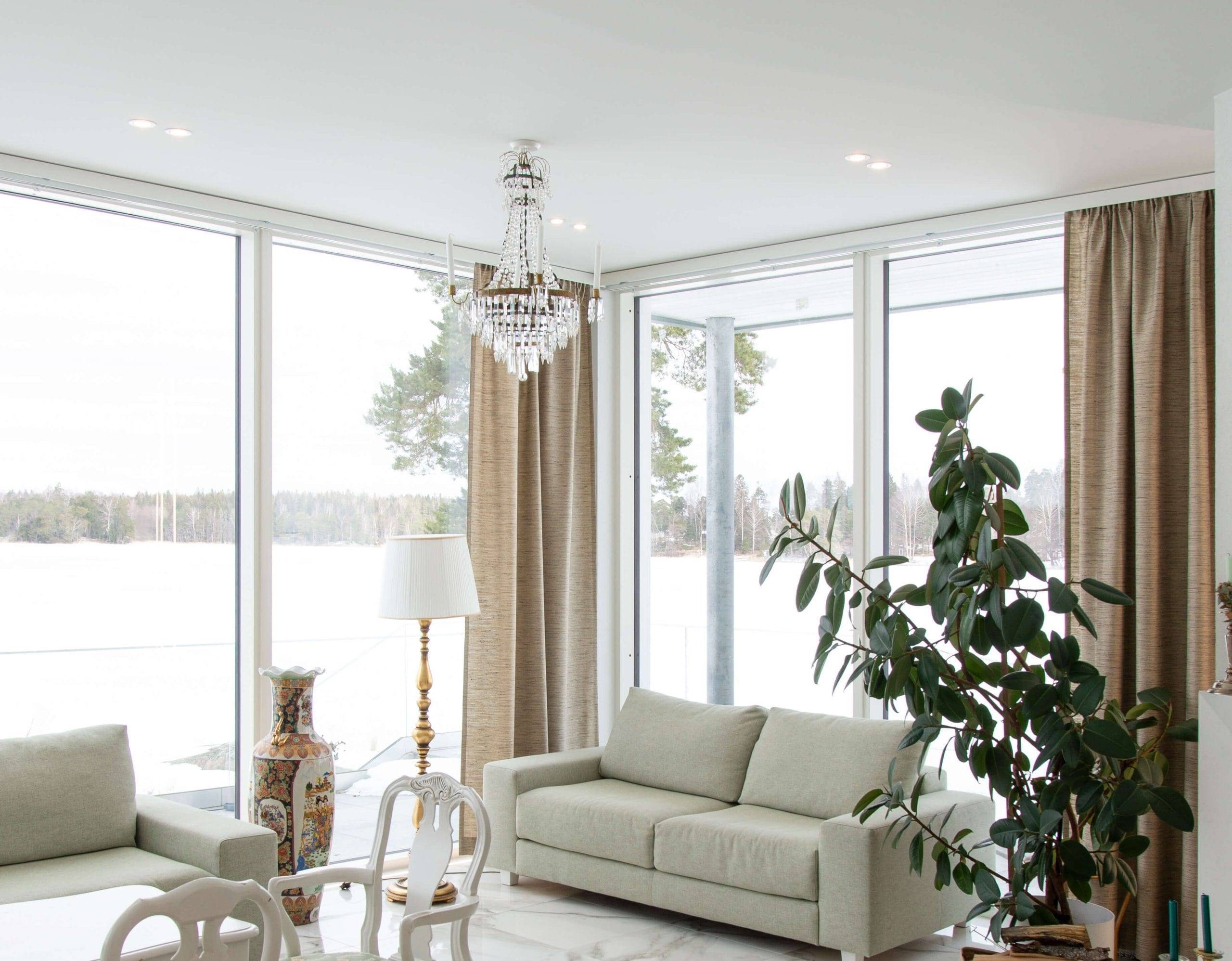 Il luminoso soggiorno, con ampie finestre che si affacciano sul paesaggio innevato, presenta un divano bianco, una grande pianta verde e un elegante lampadario con luci a LED che ne esaltano la bellezza. Le tende beige completano un vaso decorativo e una lampada da terra, mentre un tavolo da pranzo con sedie bianche brilla sullo sfondo. Dai un'occhiata all'ampia gamma di apparecchi LED di LedStore.fi e crea una soluzione di illuminazione altrettanto straordinaria per la tua casa!