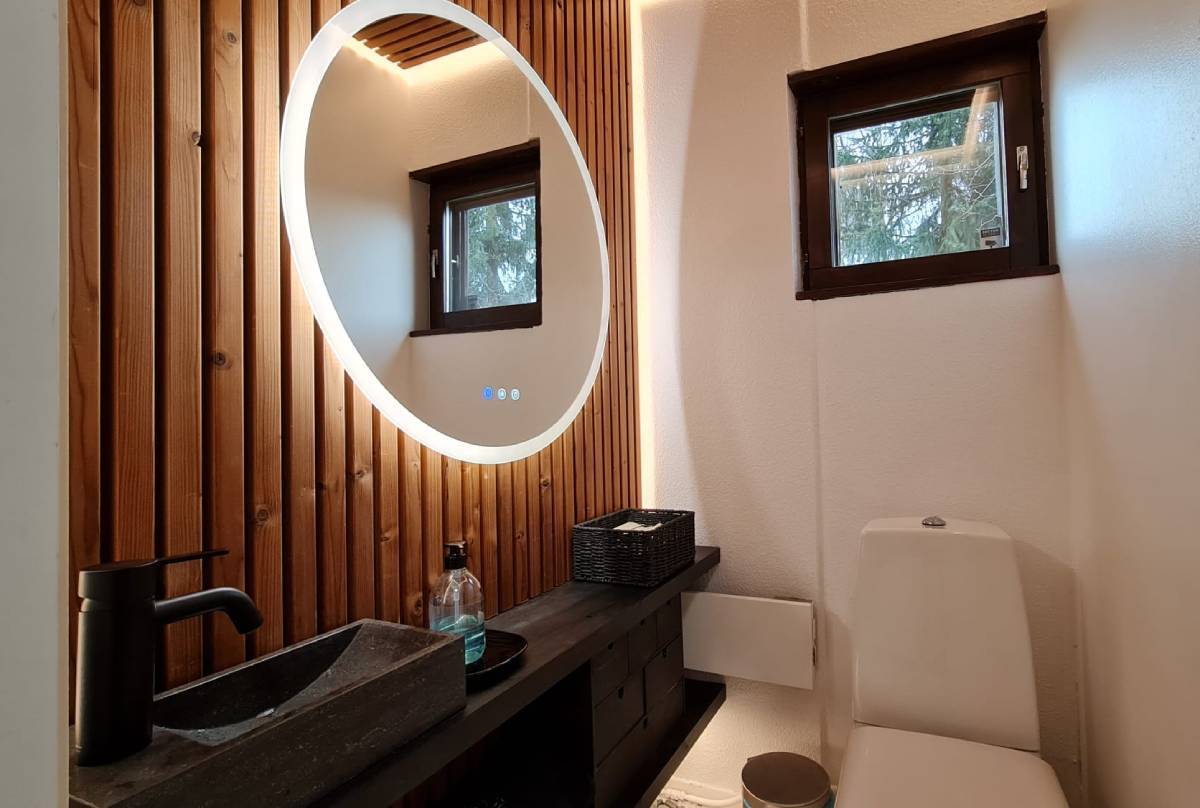 Un bagno moderno con illuminazione a LED fornita da LedStore.fi comprende uno specchio circolare a LED sopra una parete in pannelli di legno. Sotto lo specchio si trova un piano d'appoggio nero con un lavandino rettangolare, un rubinetto nero e un dispenser di sapone. Una piccola finestra si trova sopra la tavoletta del water e nelle vicinanze si trova una pratica soluzione per riporre gli oggetti.