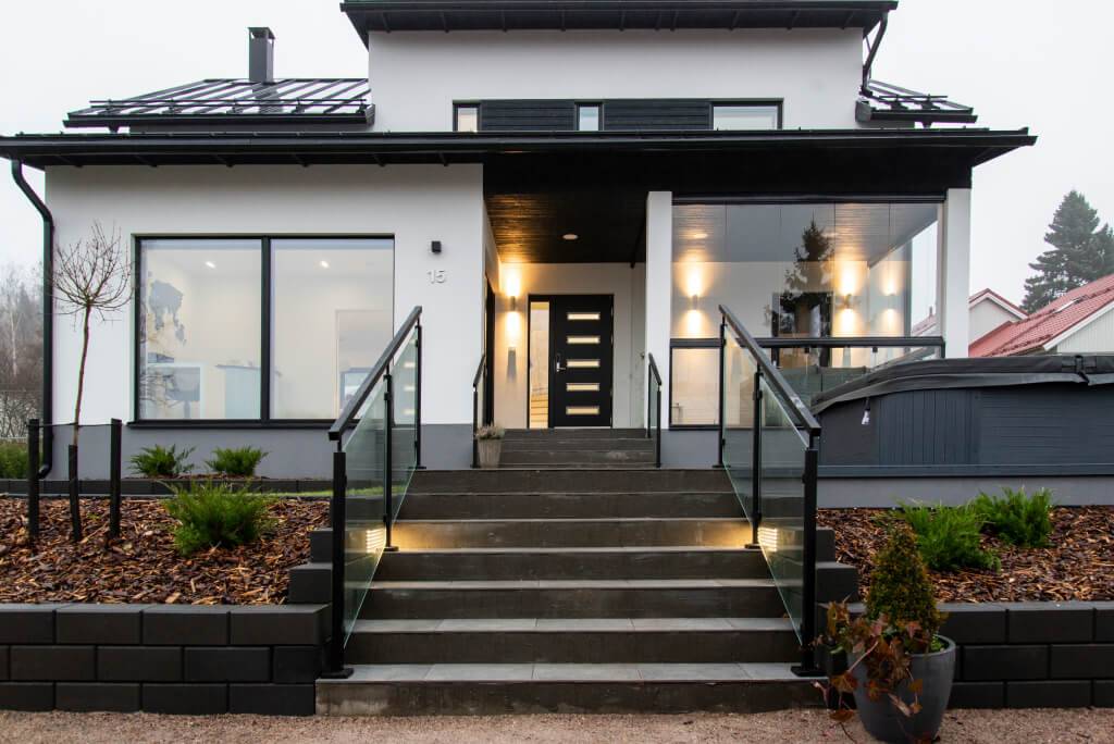 Casa moderna con facciata bianca e accenti neri; scale con balaustre in vetro e luci LED. Ampie vetrate, paesaggio minimo, arbusti e casa vicina.