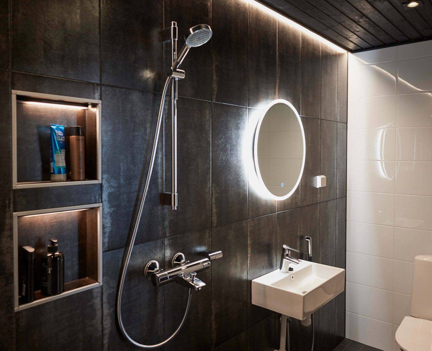 Bagno elegante con piastrelle scure, specchio retroilluminato a LED sopra il lavandino, doccia, mensole integrate e soffitto nero. Una toilette moderna completa il look elegante.