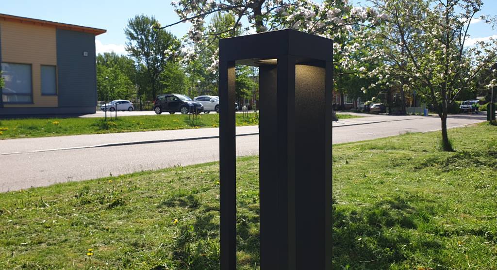 Un moderno lampione nero con luce LED lungo la strada; alberi in fiore, auto parcheggiate e un edificio con grandi finestre sullo sfondo sotto un cielo soleggiato.