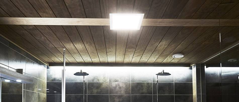 Bagno moderno con soffitto in legno, illuminazione LED quadrata, pareti piastrellate scure, due soffioni a pioggia e un'atmosfera calda grazie all'illuminazione LED soffusa.