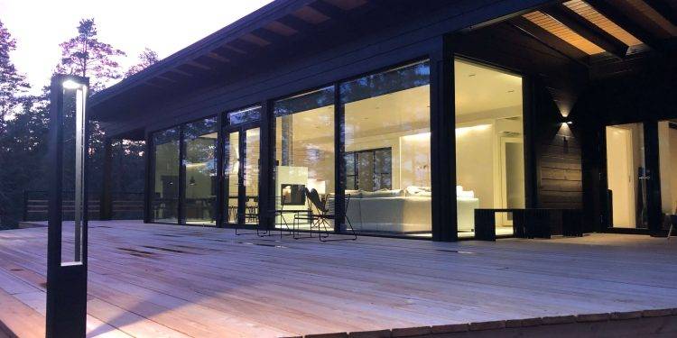 Casa moderna con ampie vetrate e spaziosa terrazza in legno, circondata da alberi. Gli interni sono luminosi e presentano un design minimalista con toni dipinti a mano. Le luci esterne a LED di LedStore.fi aggiungono una calda luce al crepuscolo serale.