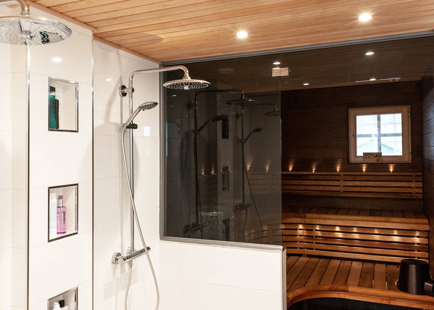 Bagno moderno con due docce a pioggia, parete in vetro, scaffale per articoli da toilette incorporato, sauna in legno adiacente, illuminazione d'atmosfera alle finestre e illuminazione a LED.