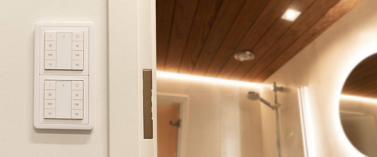 Bagno con pannelli a soffitto in legno e illuminazione soffusa a LED. C'è anche una comoda cabina doccia con soffione a pioggia. Su una parete vicina si trovano due moderni interruttori per la luce, controllati da tastiere. L'insieme elegante è coronato da uno specchio circolare illuminato a LED.